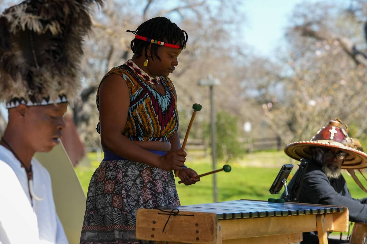 Outdoor musicians perform traditional percussion, with a standing marimba player in patterned dress and seated performers in ceremonial attire.