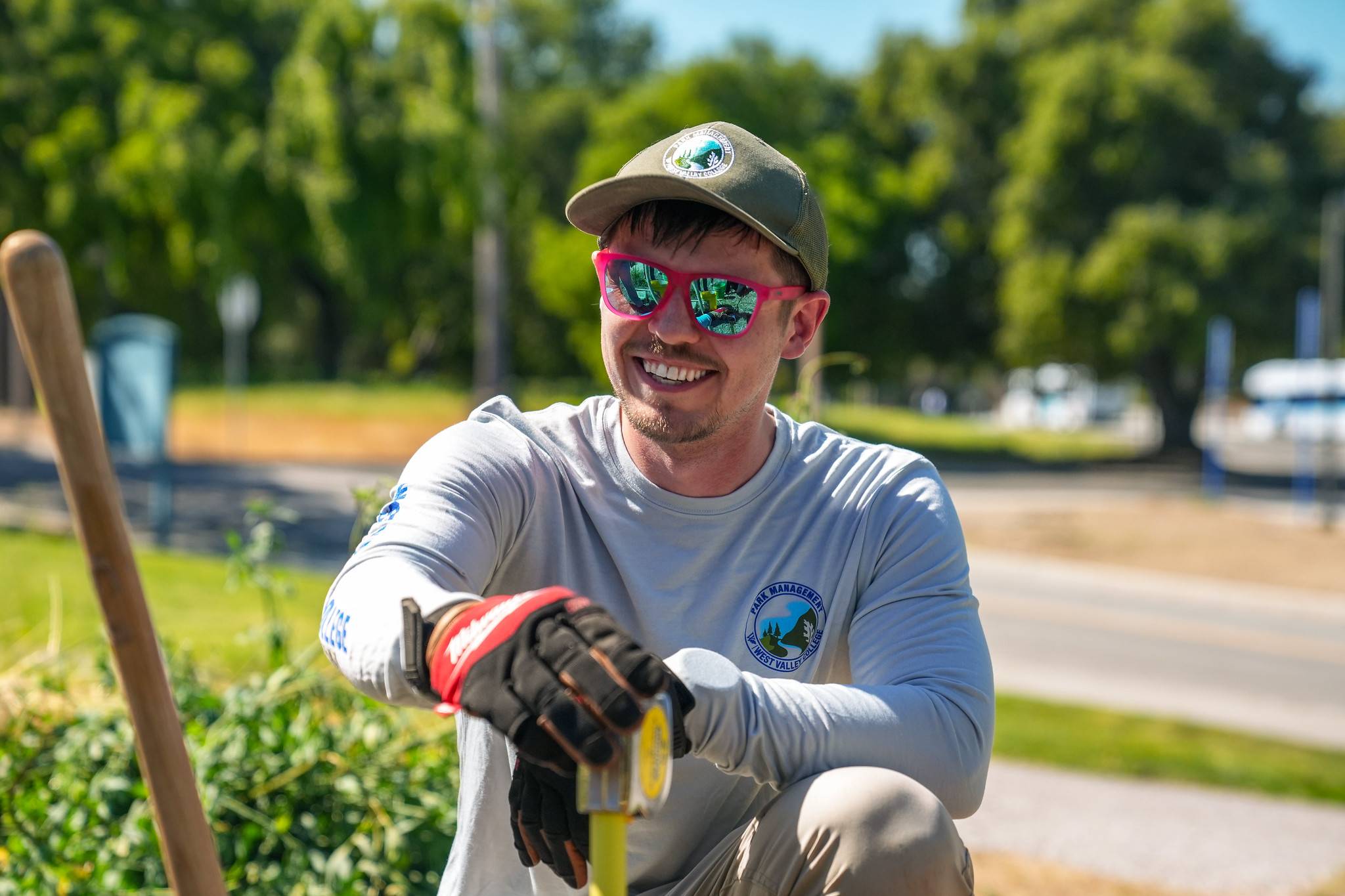 parks management student sitting down in the field wearing pink polarized sunglasses, a light green dad hat and field gloves. He is holding a shovel